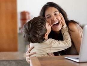 Niño dando un beso en el cachete a su madre sonriente. La mamá está sentada en frente de un ordenador portátil