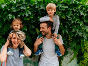 Estilos de crianza Mamá y papá al aire libre, alzando cada uno a sus hijos sobre sus hombros y sonriendo.
