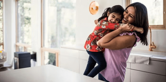 ¿Cuáles son los estilos de crianza? Mamá alzando y abrazando a su hija en la cocina, ambas sonriendo felices.